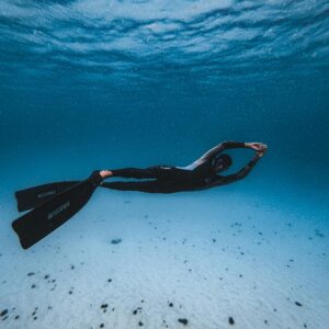 A man in a black wetsuit swimming underwater in clear blue water, showcasing diving and exploration.